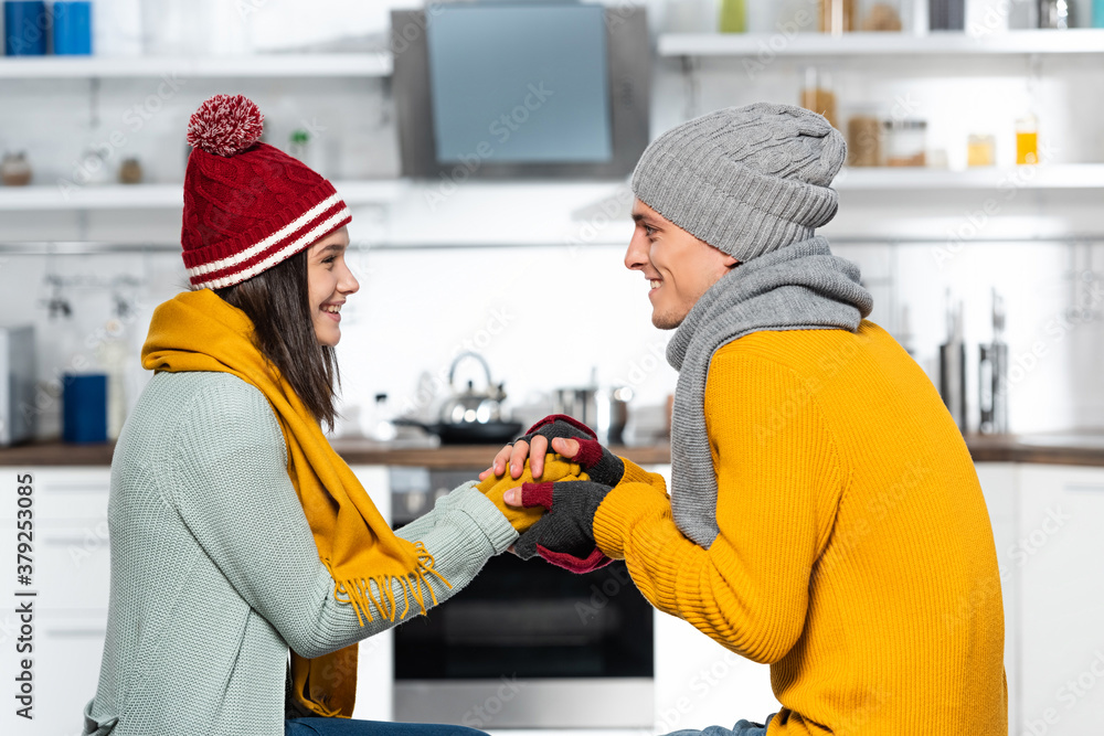 side view of man in knitted hat warming hands of freezing girlfriend in kitchen side view of man in knitted hat warming hands of freezing girlfriend in kitchen