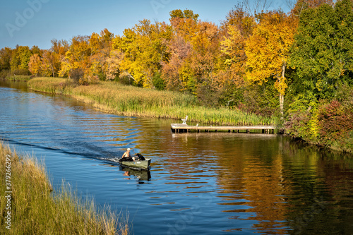 The beautiful Mississippi River in Bemidji, Minnesota with a small boat carrying a man and two black lab dogs on the blue water and autumn colors in the trees along the shoreline.