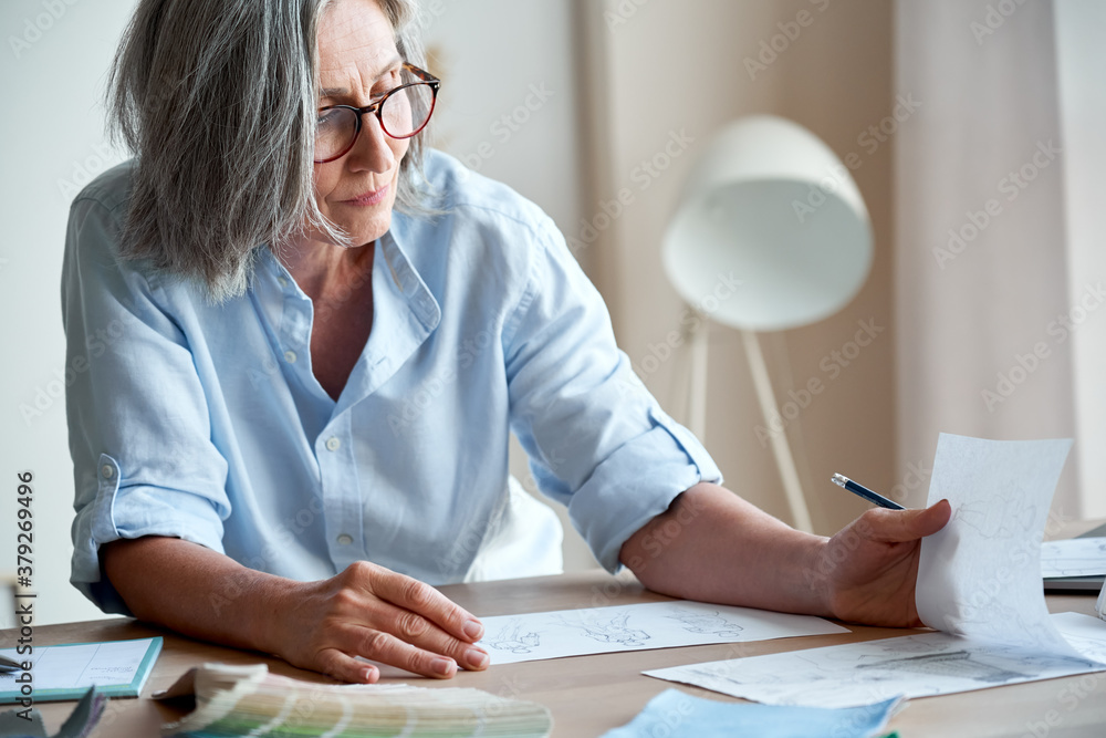 Mature old adult elegant woman fashion designer drawing creative sketches on table. Beautiful sophisticated middle aged grey-haired lady entrepreneur creating new fashion design cloth in atelier. Mature old adult elegant woman fashion designer drawing creative sketches on table. Beautiful sophisticated middle aged grey-haired lady entrepreneur creating new fashion design cloth in atelier.