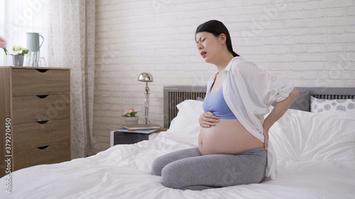 Foto full length korean pregnant woman kneeling on bed is placing one hand on stomach and other on her back, massaging and moaning in pain