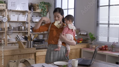 busy asian career mom holding baby is gripping the toast in mouth, carrying her bag and coat and leaving home to rush to work.