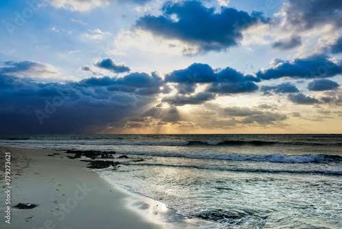 The waves of the Caribbean Sea roll ashore. The rays of the rising sun penetrate the clouds  paint the sky in golden tones. There are no people. Only footprints in the sand of the beach. Mexico.