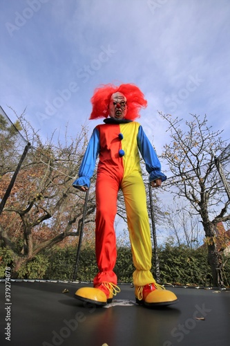 Halloween party .Scary aggressive clown with red hair jumping in a trampoline on autumn trees and blue sky background. Creepy clown costume. Festival and carnival. 