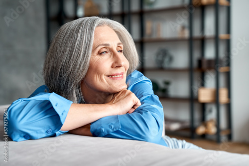 Happy relaxed mature old woman resting dreaming sitting on couch at home. Smiling mid aged woman relaxing. Peaceful serene grey-haired lady feeling peace of mind enjoying lounge on sofa and thinking.