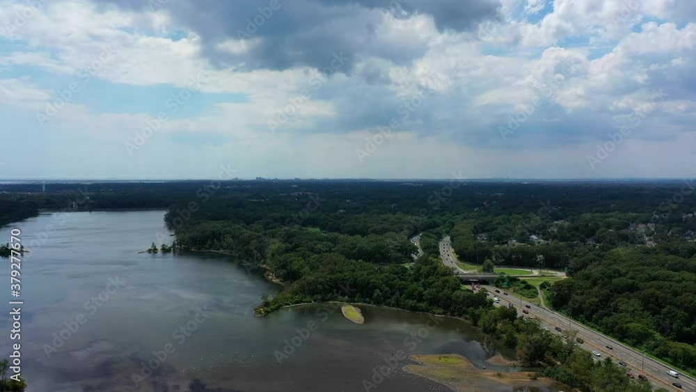 An aerial view over a reflective lake. It is a beautiful, sunny day with white clouds. The camera truck right and pan left. There are lots of green trees and a highway with some cars driving by. An aerial view over a reflective lake. It is a beautiful, sunny day with white clouds. The camera truck right and pan left. There are lots of green trees and a highway with some cars driving by.