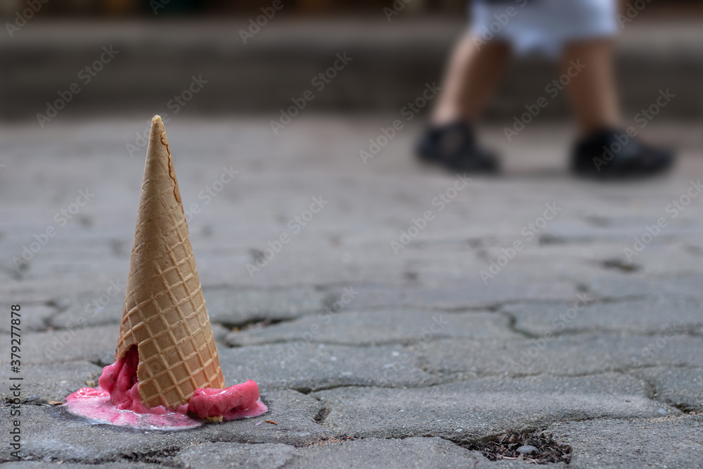 Close up image of fallen pink strawberry sorbet ice cream with cracking cone on the old cement ground and blurred children legs background. Selective focus and copy space. Close up image of fallen pink strawberry sorbet ice cream with cracking cone on the old cement ground and blurred children legs background. Selective focus and copy space.