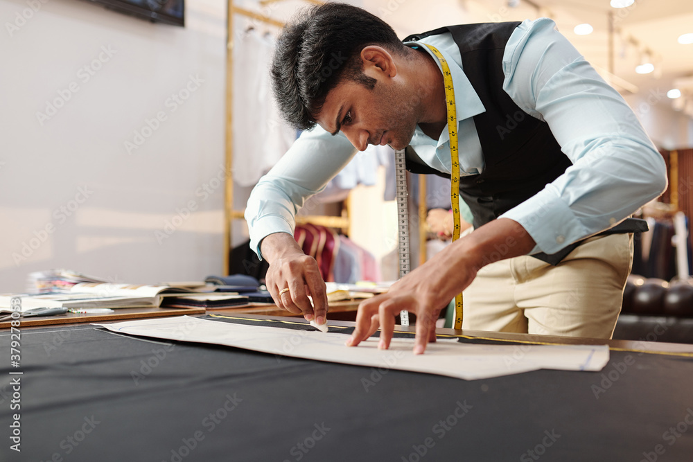 Concentrated young Indian tailor tracing sewing pattern on black fabric Concentrated young Indian tailor tracing sewing pattern on black fabric