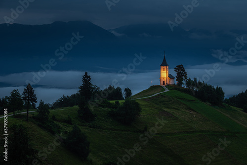 Jamnik, Slovenia - Blue hour at Jamnik with illuminated St. Primoz hilltop church on a foggy summer dawn. Julian Alps at background