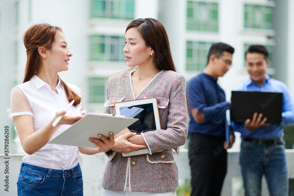 Serious young Asian businesswoman looking at her colleague with clipboard explaining her creative ideas Serious young Asian businesswoman looking at her colleague with clipboard explaining her creative ideas