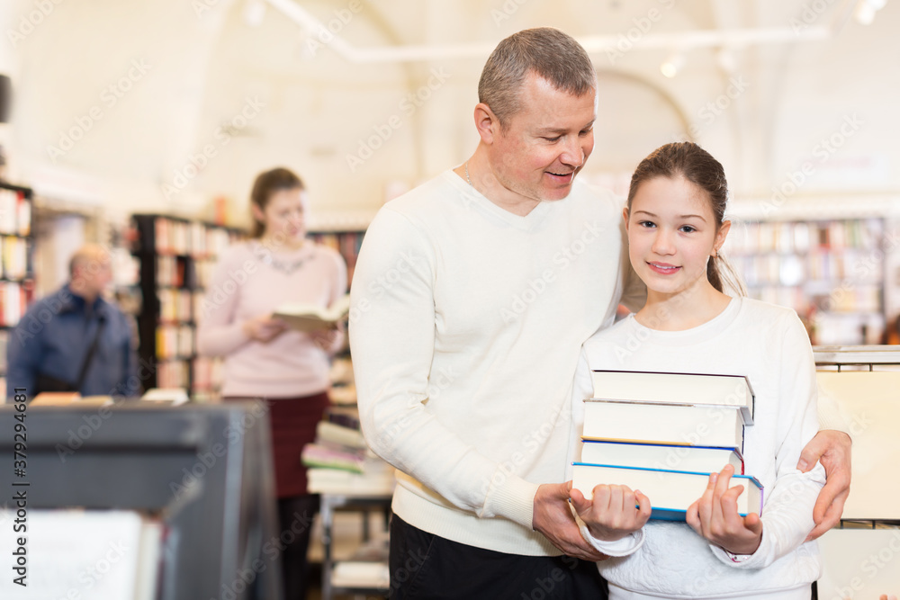 Obraz premium Intelligent preteen girl with father standing with stack of books chosen at bookshop