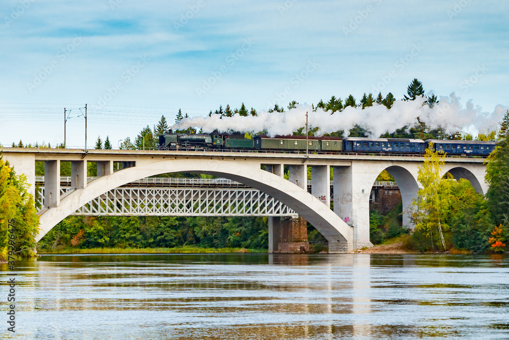 Naklejka premium Kouvola, Finland - 18 September 2020: Autumn landscape of bridge with moving old steam passenger train Ukko-Pekka and Kymijoki river waters in Finland, Kouvola, Koria