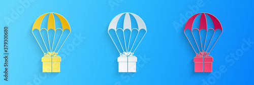 Paper cut Gift box flying on parachute icon isolated on blue background. Delivery service, air shipping concept, bonus concept. Paper art style. Vector.