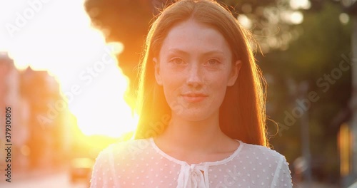 Beautiful Ginger Woman is opening her Blue Eyes while looking at Camera. Attractive girl with nice Freckles smiling Charmingly while walking on warmy Evening at Red Orange Sunset. Natural Beauty. photo