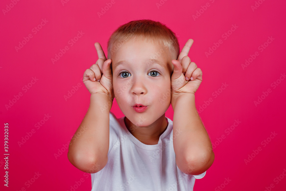 A portrait of a shocked blond boy in white casual clothes stands on a pink background, looking at the camera and pointing up with his index fingers. The surprised boy points to the copy space. A portrait of a shocked blond boy in white casual clothes stands on a pink background, looking at the camera and pointing up with his index fingers. The surprised boy points to the copy space.