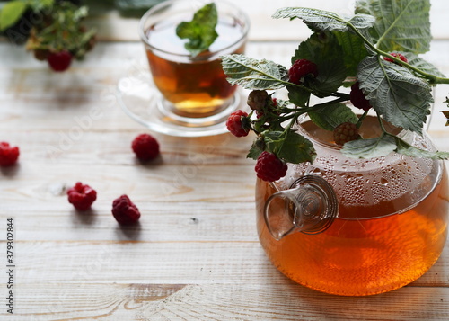 Useful properties of medicinal tea with raspberries.Branch with raspberries on a glass teapot with tea on a light background.