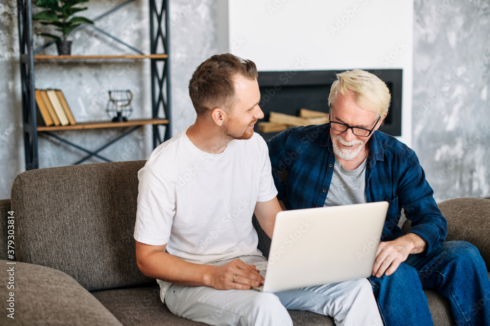 Young adult son teaches old senior father using laptop sit on sofa, two men diverse generation spend time online with computer together at home Young adult son teaches old senior father using laptop sit on sofa, two men diverse generation spend time online with computer together at home