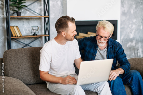 Young adult son teaches old senior father using laptop sit on sofa, two men diverse generation spend time online with computer together at home