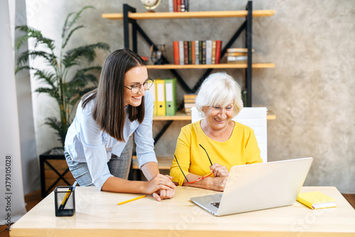 Two female colleagues diverse generation discuss about plans and business tasks at the office, elderly lady sits at the desk and using laptop, a young woman coworker stands near