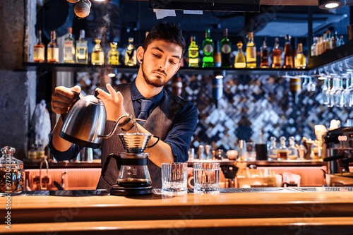 In a popular cafe with soft, yellow evening light, a professional barista pouring hot water from a metal kettle into ground coffee beans. An alternative way to make coffee. Coffee filter