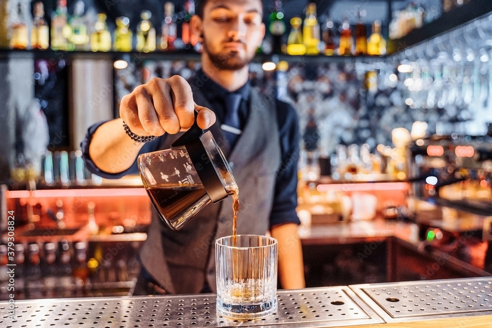 Close up of a young caucasian bearded male barista pouring coffee into Kalita Wave Dripper coffee filter. Brewing liquid after measuring coffee ground on digital scale in shop. Wearing dark uniform