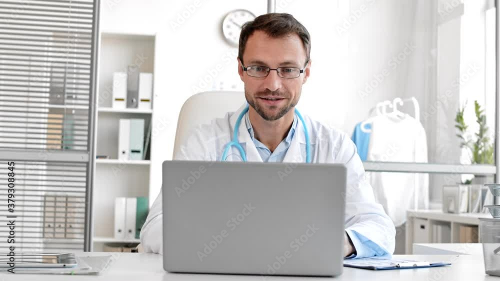 Medium shot of middle-aged Caucasian man wearing medical gown and glasses is sitting at desk in the office and talking to patient by video link Medium shot of middle-aged Caucasian man wearing medical gown and glasses is sitting at desk in the office and talking to patient by video link