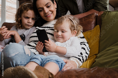 Mother and two daughters watching funny videos on smartphone sitting on sofa in modern lounge.