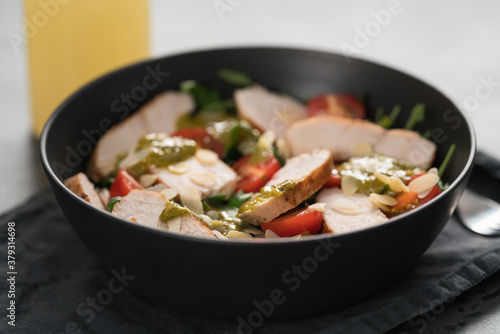 Salad with chicken, tomatoes, arugula and pesto in black bowl on concrete background