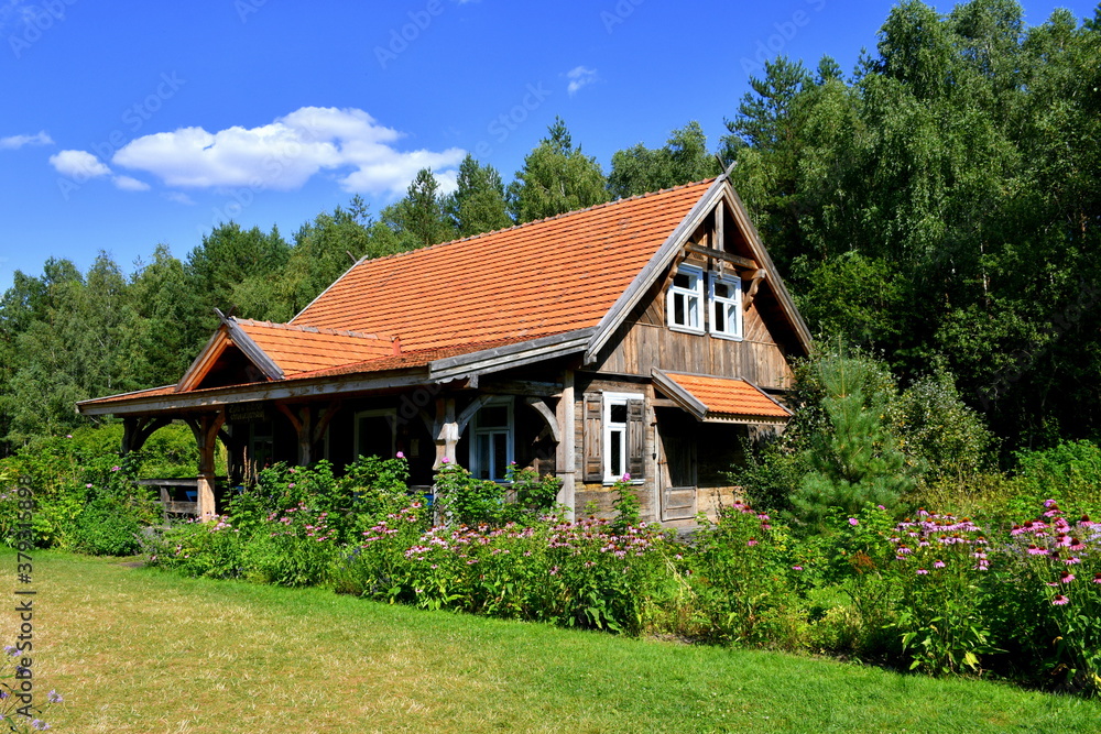 Close up on old rural house with an angled roof and its walls made entirely out of logs, planks, and boards with a neatly maintained garden seen in front of it next to a grass covered lawn in Poland
