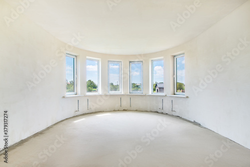 The unfinished living room in a new residential house with the white plastered curved walls and a row of the window frames