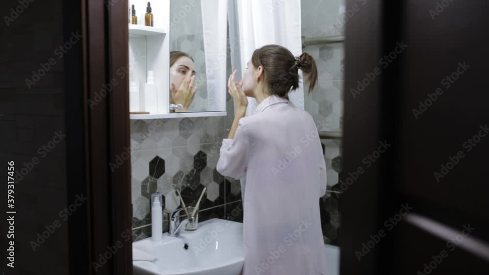 Back view of smiling woman in domestic gown standing in bathroom in front of mirror and applying facial mask while pampering herself at weekend and doing beauty procedure at home Back view of smiling woman in domestic gown standing in bathroom in front of mirror and applying facial mask while pampering herself at weekend and doing beauty procedure at home