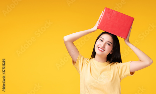 Friendly young woman holding red gift box in her hands while looking at camera over isolated against yellow background. Excited teenager received surprise gift for celebrate or anniversary.