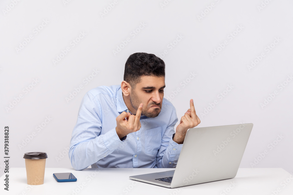 Annoyed businessman sitting office workplace, showing middle finger to laptop screen, expressing hate to job