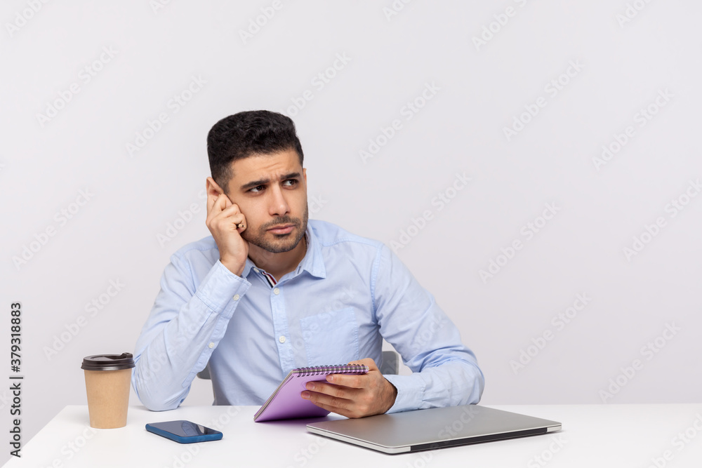 Thoughtful journalist or businessman sitting in office, holding paper notebook and leaning on hand dreaming, thinking Thoughtful journalist or businessman sitting in office, holding paper notebook and leaning on hand dreaming, thinking