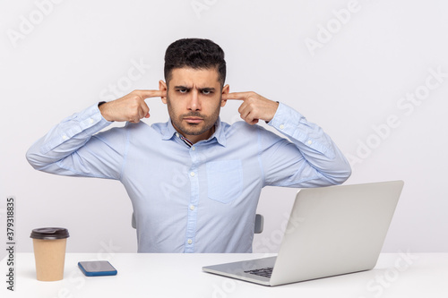 Can't hear this anymore! Annoyed man employee sitting at workplace, covering ears and frowning angry displeased