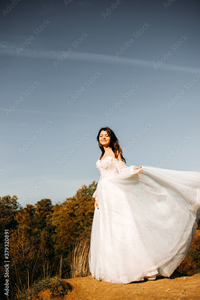 Gorgeous bride in a white dress posing on a hill against the sky.