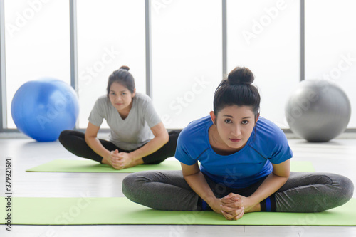 Two middle-aged Asian women doing yoga sitting on a rubber mat in a gym.