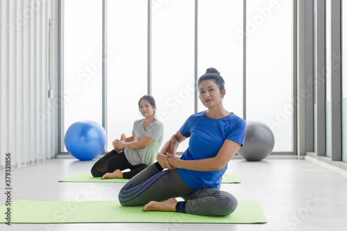 Two middle-aged Asian women doing yoga sitting on a rubber mat in a gym.