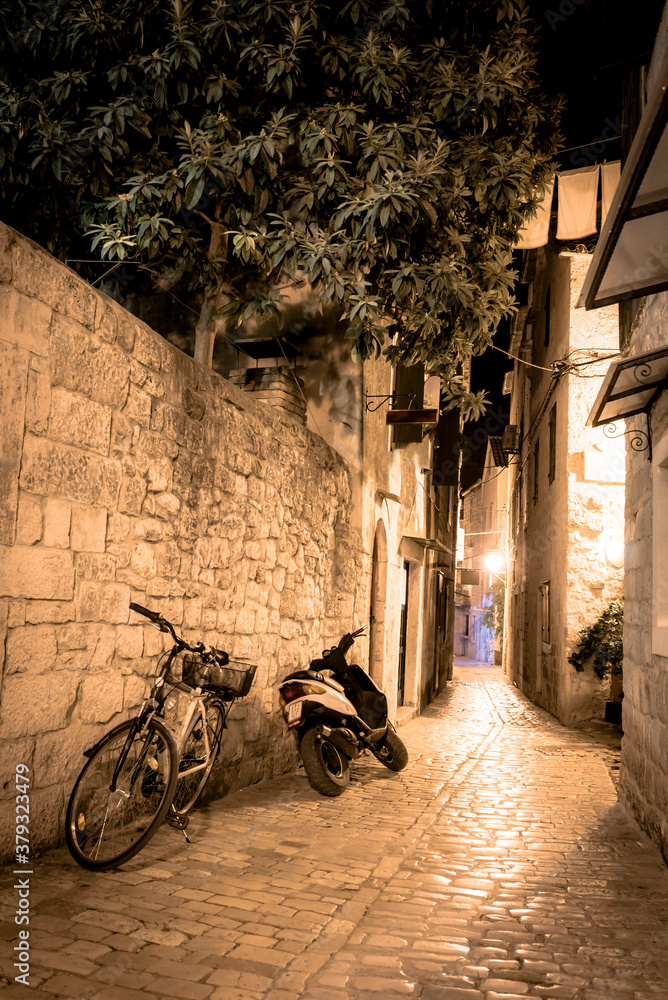 A narrow deserted street with bike and motorcycle in the medieval historical part of Trogir, Croatia lit by lanterns at night