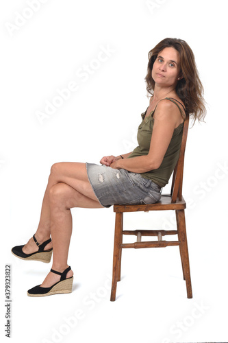 side view of a serious woman in denim skirt sitting on a chair and looking at camera on white background, legs crossed