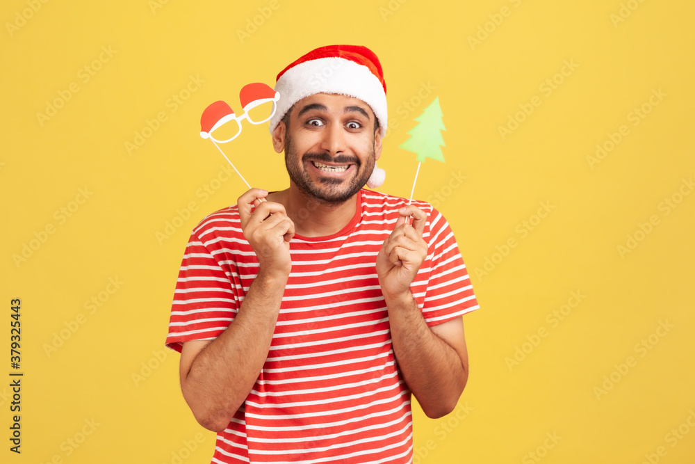 Extremely happy man in red t-shirt and santa claus hat having fun holding paper goggles and christmas tree on stick, celebrating christmas eve. Indoor studio shot isolated on yellow background Extremely happy man in red t-shirt and santa claus hat having fun holding paper goggles and christmas tree on stick, celebrating christmas eve. Indoor studio shot isolated on yellow background