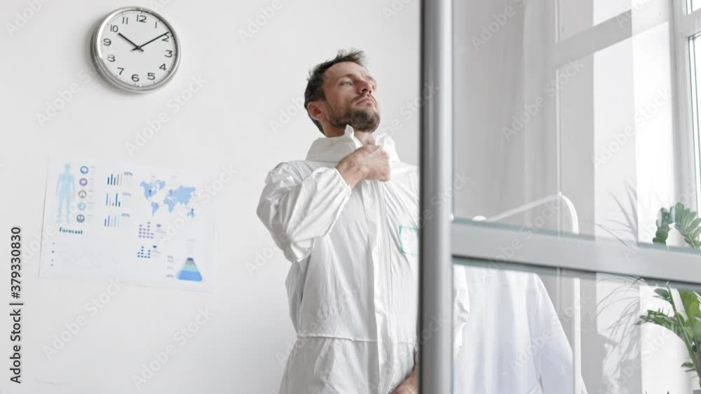 Panning of middle-aged Caucasian male virologist is standing in the office, zipping up white protective coverall and going away Panning of middle-aged Caucasian male virologist is standing in the office, zipping up white protective coverall and going away
