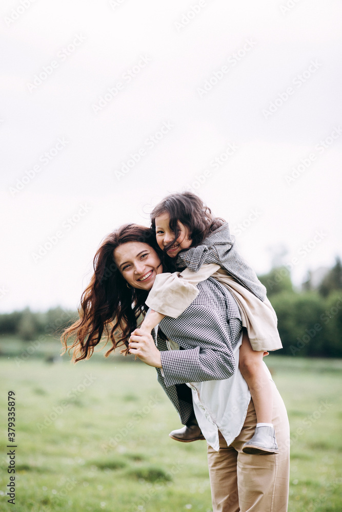 Stylish mother and daughter have fun outdoors in a field with green grass by the river when it rains