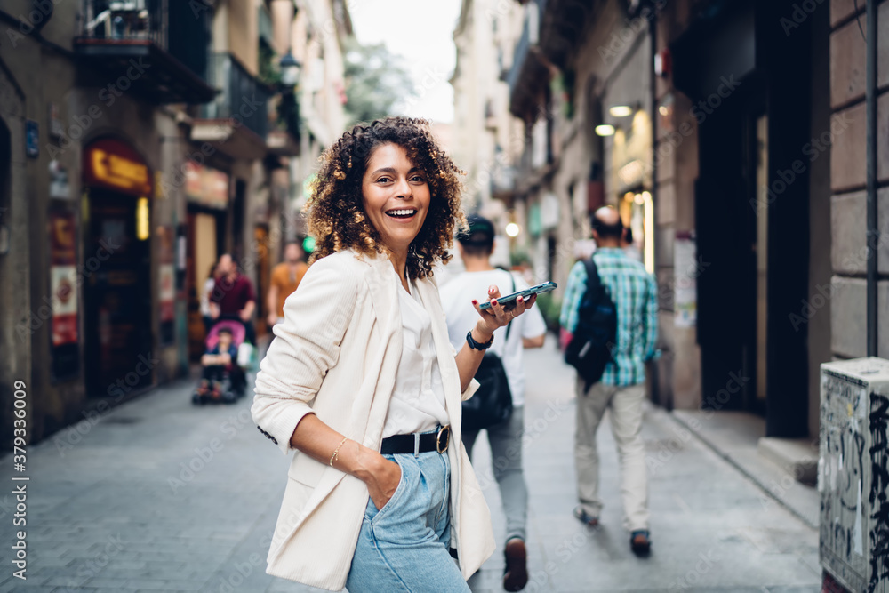 Positive young ethnic lady standing with smartphone on sidewalk in aged town