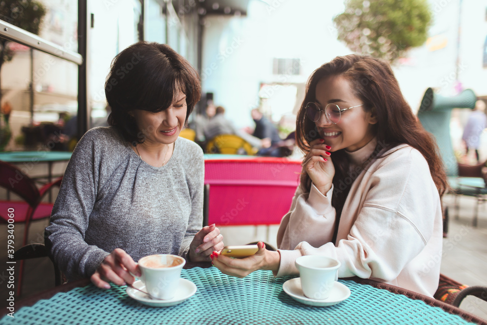 Mature mother and her young daughter sit together in cafe or restaurant. Girl hold smartphone and showing something on screen to her mom. Mature woman watch it. Drinking tea or coffee. Mature mother and her young daughter sit together in cafe or restaurant. Girl hold smartphone and showing something on screen to her mom. Mature woman watch it. Drinking tea or coffee.