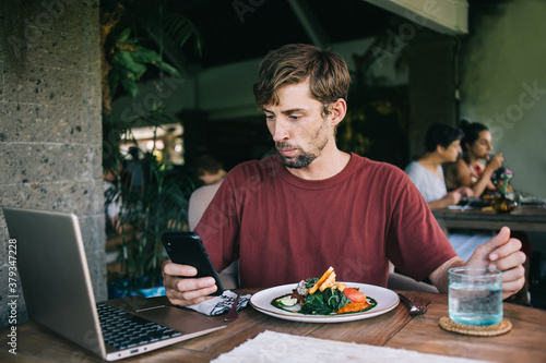 Serious caucasian male freelancer sitting in cafe interior during break for dinner checking income messages on smartphone, pensive hipster guy checking banking account making payment for food