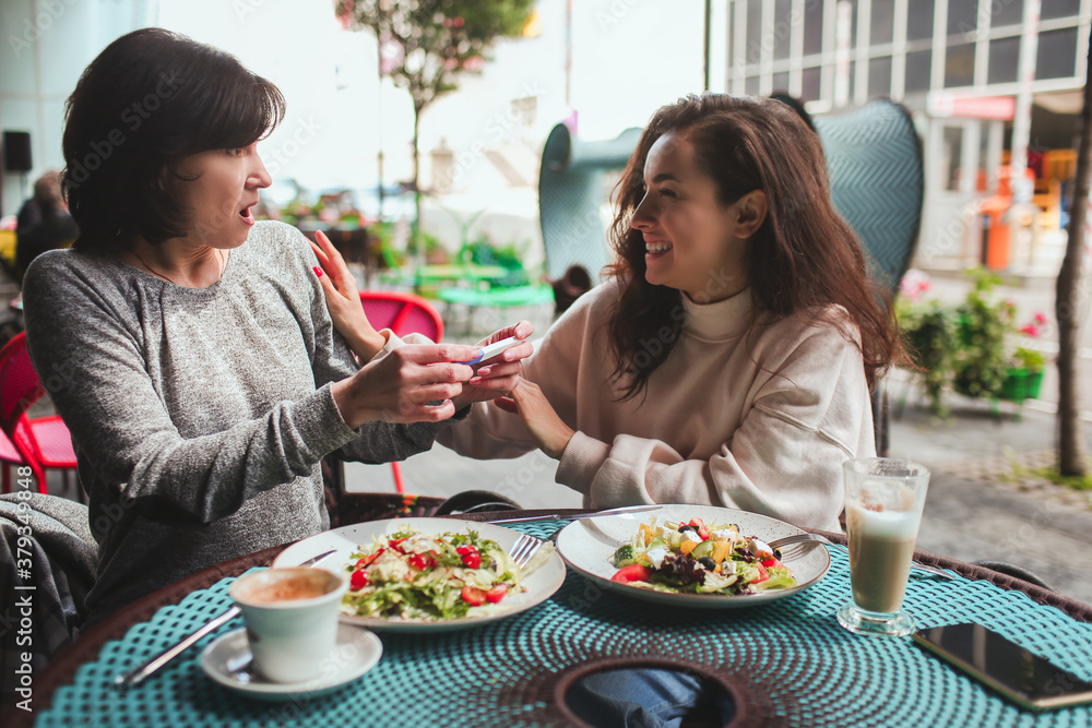 Mature mother and her young daughter sit together in cafe or restaurant. Shocked mature woman hearing about her daughter's pregnancy. Both of them happy and rejoicing.