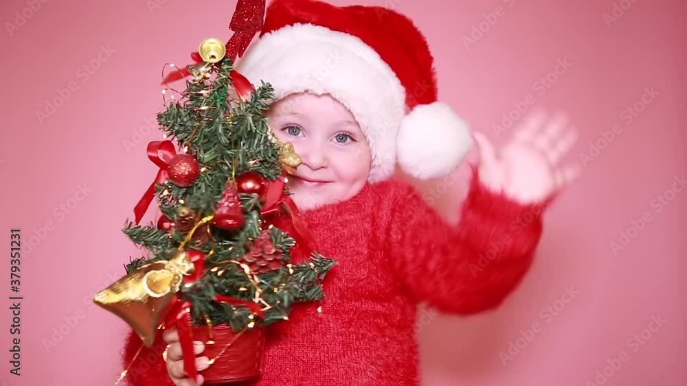 A little girl is sitting in a Santa hat, holding a Christmas tree, smiling funny, waving, greeting. Isolate on a pink background A little girl is sitting in a Santa hat, holding a Christmas tree, smiling funny, waving, greeting. Isolate on a pink background