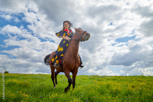 Beautiful gypsy girl rides a horse in field with green glass in summer day and blue sky with white clouds. Model in ethnic dress posing with farm animal