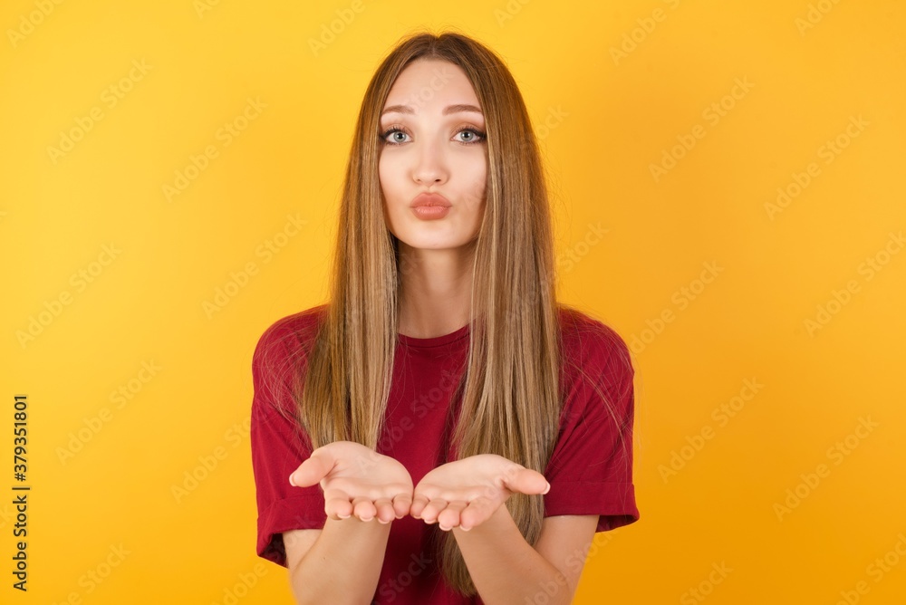 Beautiful Young beautiful caucasian girl wearing red t-shirt over isolated yellow background sending blow kiss with pout lips and holding palms to send air kiss. Beautiful Young beautiful caucasian girl wearing red t-shirt over isolated yellow background sending blow kiss with pout lips and holding palms to send air kiss.