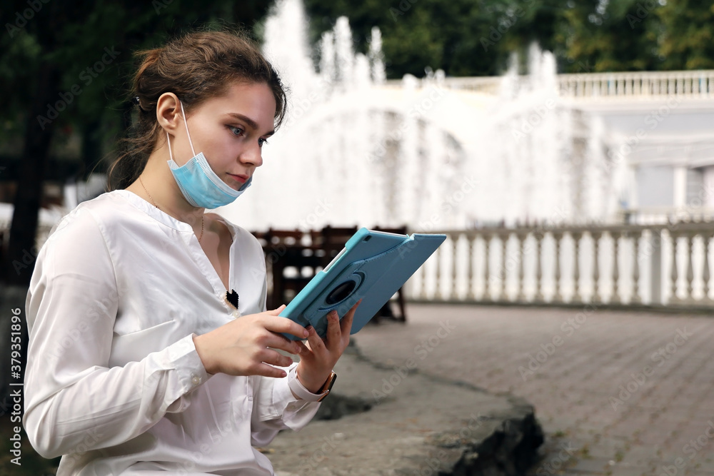 Obraz premium Young woman student in medical facial mask sits in quiet, deserted place in the park against the background of fountains. She looks at the tablet intently